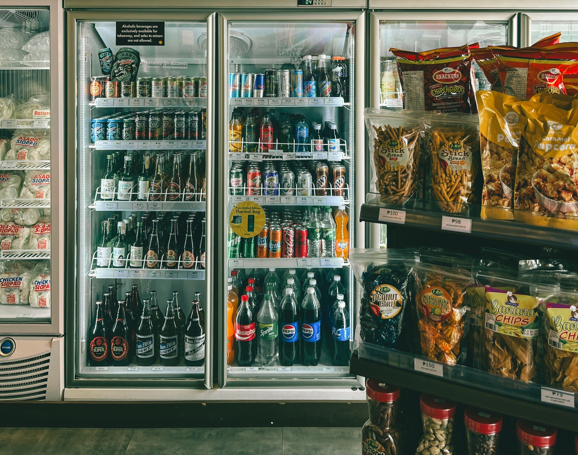 Refrigerated vending display with bottled drinks, snacks, and chips, priced labels visible inside glass doors.